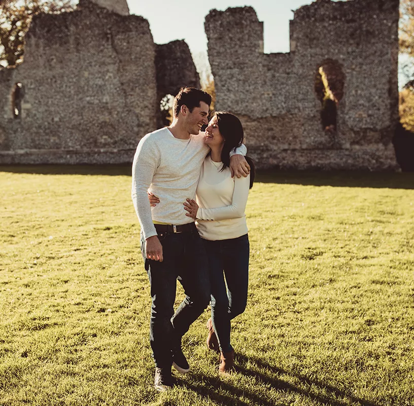 Happy couple with white jumpers and blue jeans walking with arms around each other smiling and looking at each other with ruins in the background.
