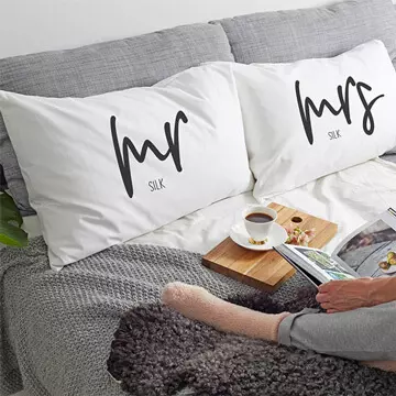 White Mr & Mrs pillow set sitting on a grey bed and person reading a book.