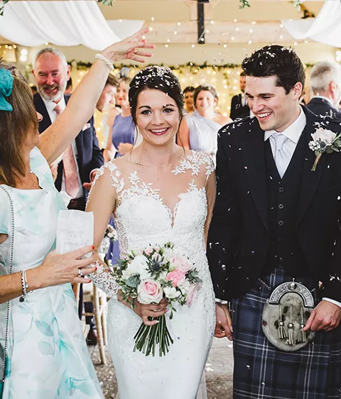 A young couple in their 20s holding hands and smiling after getting married walking down the aisle with white drapes and confetti being thrown. Brunette lady wearing a beautiful white dress holding a bouquet of flowers. Clean shaved man wearing a blue tartan kilt.