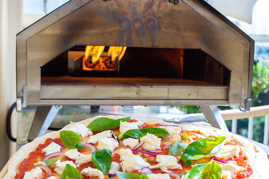 A freshly baked pizza with basil and cheese in front of an Ooni pizza oven with flames visible inside.