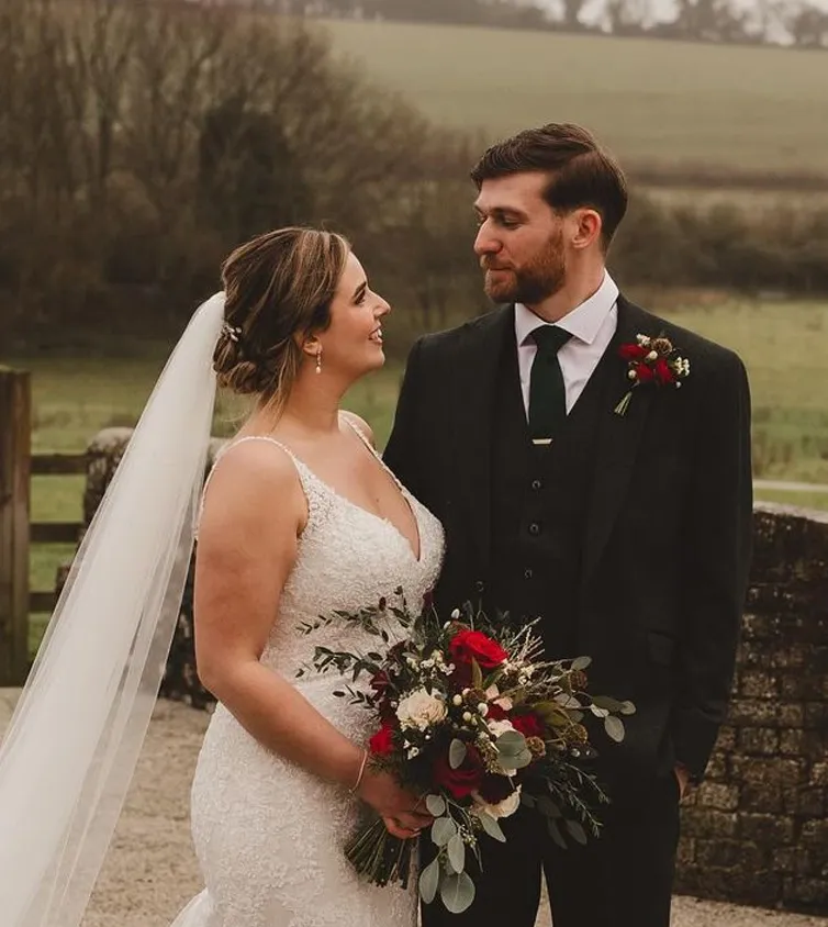 A man wearing a green tweed suit with a dark green tie and his wife in a white wedding dress holding red and white roses