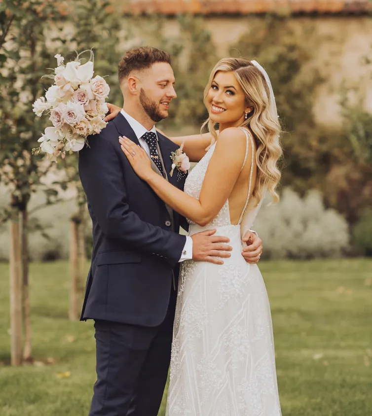 Bearded Man in a suit and Blonde woman in a wedding dress hugging with a green background.