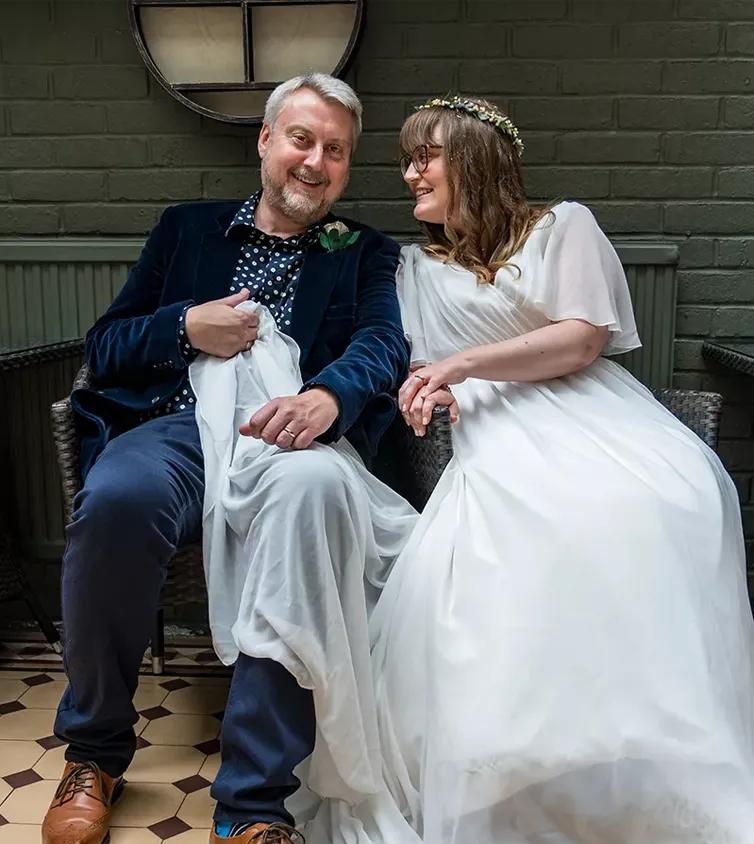 White hair man just married sitting on chairs holding the brides wedding dress smiling.